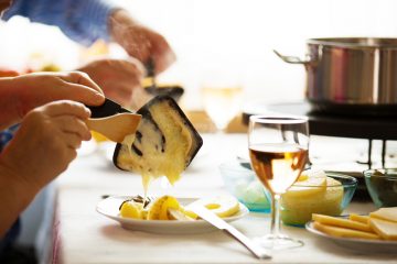 Une personne se sert le fromage fondu de sa raclette. Un verre de vin blanc sur la table.