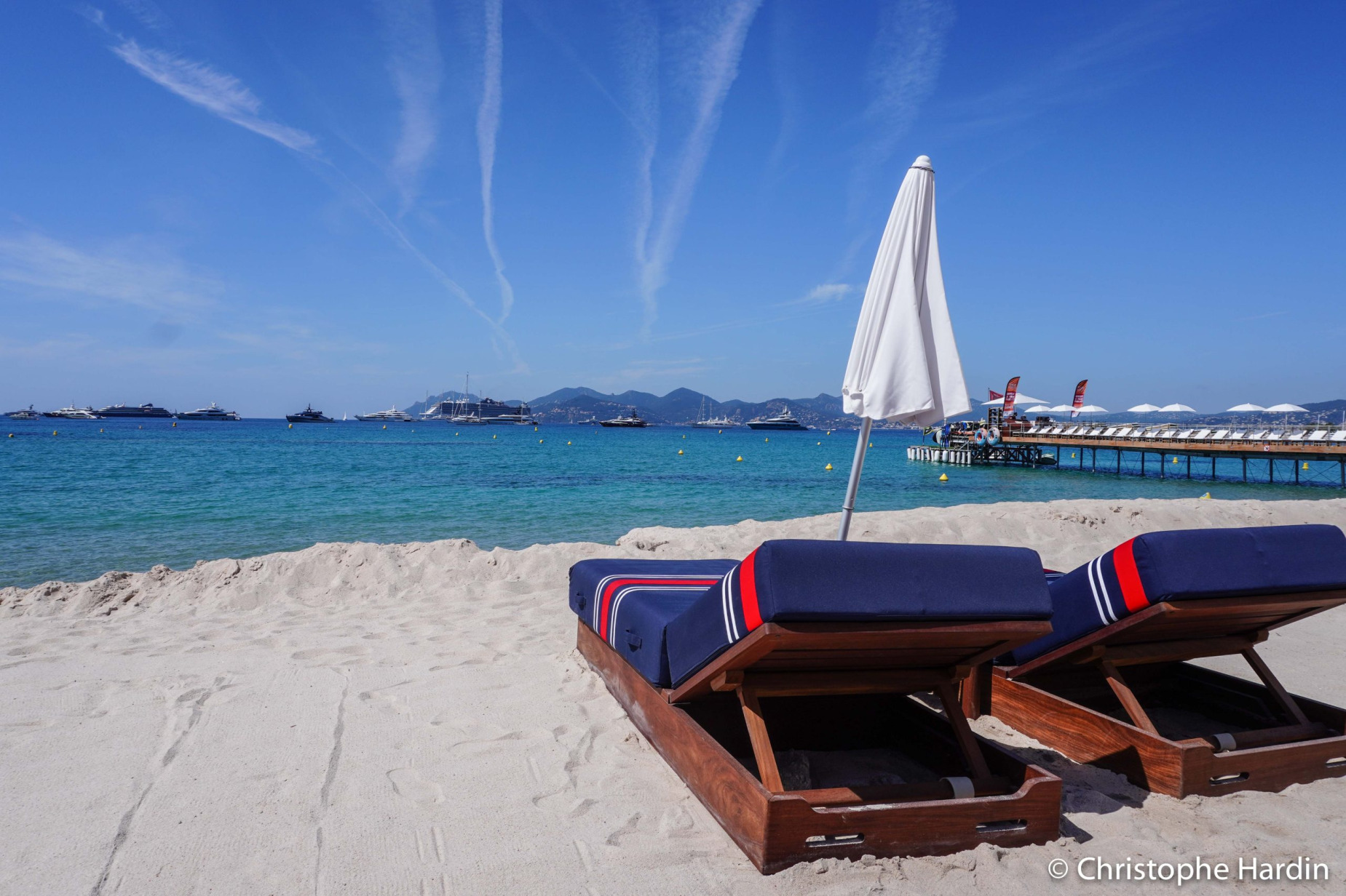 Deux transats aux couleurs de la compagnie aérienne Air France bleu blanc rouge et un parasol blanc fermé sur la plage de Cannes avec la mer et le ciel en arrière plan.