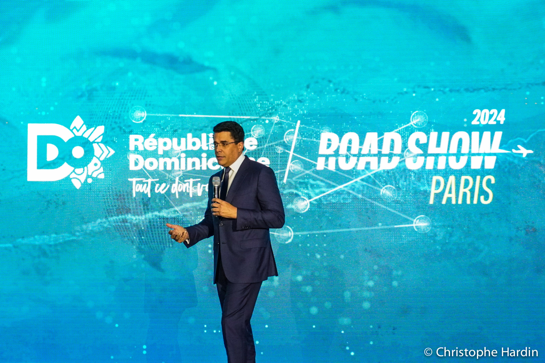 David Collabo en costume bleu marine devant un écran bleu turquoise sur lequel est inscrit : République Dominicaine, tout ce dont vous avez besoin et Roadshow Paris 2024.