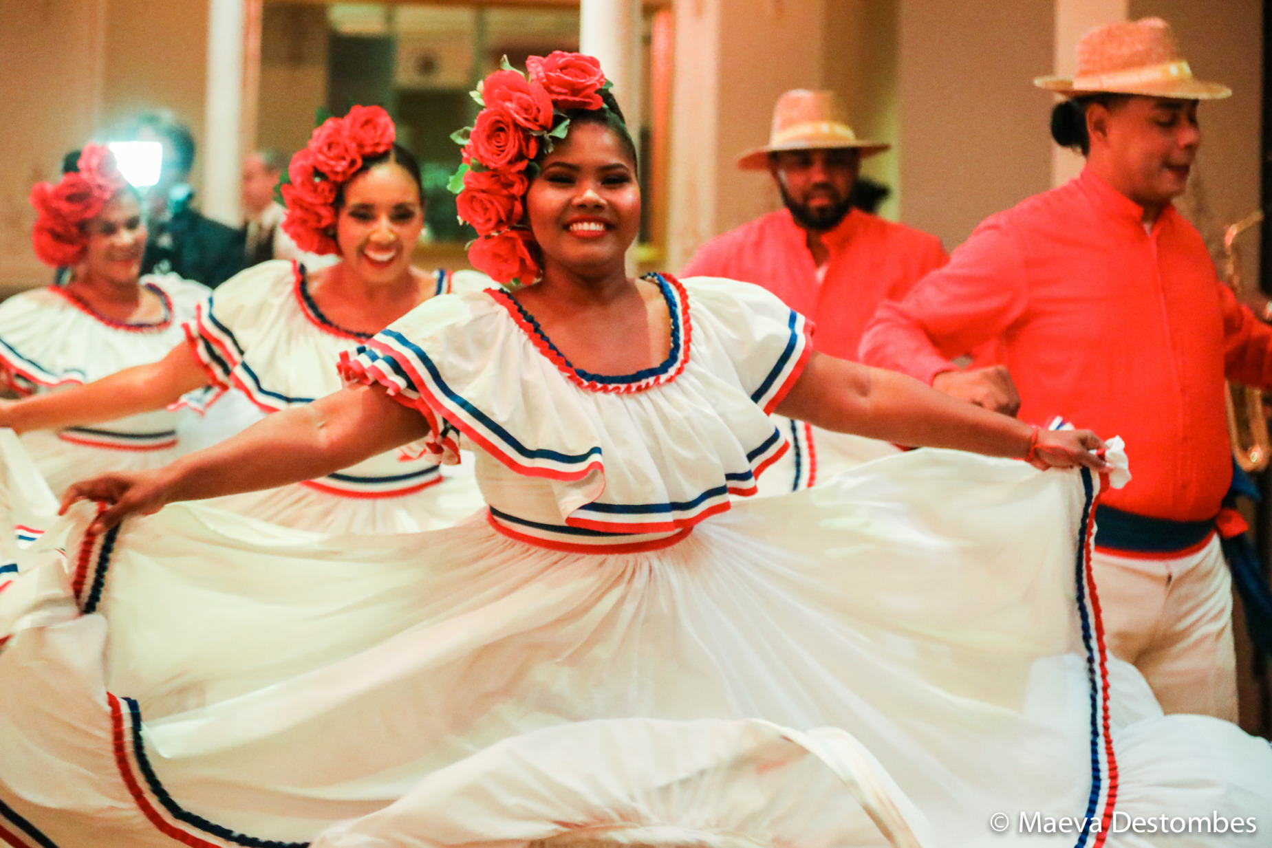 Des danseuses en robe blanche bordée d'un liseré bleu blanc rouge, portant des couronnes de fleurs rouges, dansent.