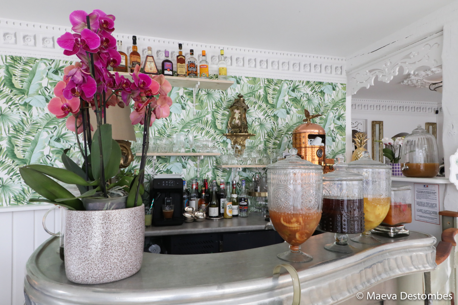 Sur le comptoir du bar, des bonbonnes de cocktail colorées s’alignent avec élégance, tandis qu’un grand samovar en cuivre et un pot d’orchidées aux fleurs éclatantes composent une décoration raffinée et chaleureuse, invitant à la découverte et à la convivialité.